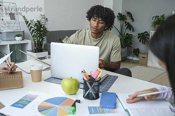 Curly haired man sitting and studying on laptop with friend at home