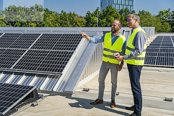 Two workers in safety vests discuss on a rooftop with solar panels while holding a tablet computer