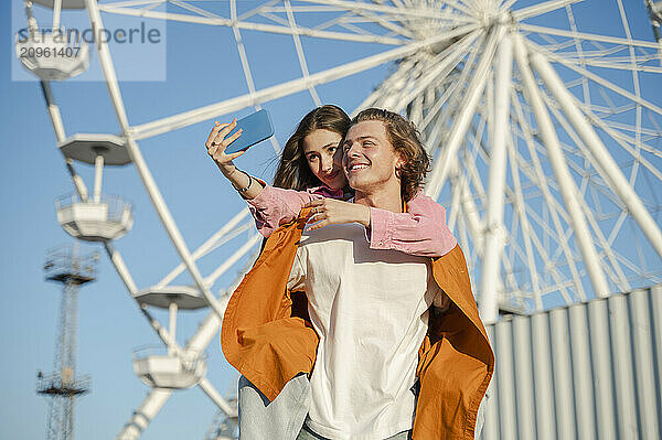 Smiling couple clicking selfie in front of ferris wheel at amusement park