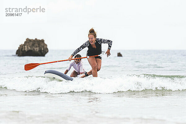 Playful woman surfing on paddle board with granddaughter at sea