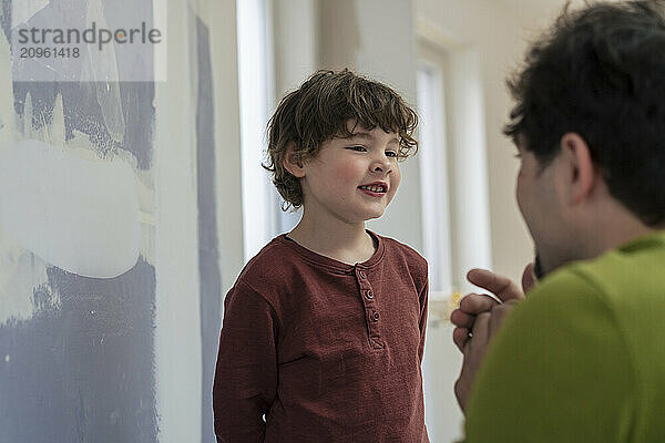 Smiling boy standing near incomplete painted wall and talking with father at home