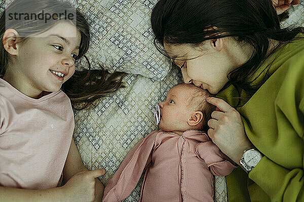 Mother kissing newborn baby girl lying besides sister on bed