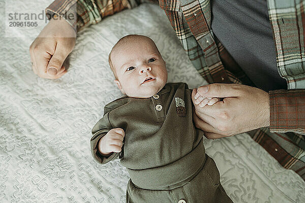 Father holding hand of baby boy lying on bed at home