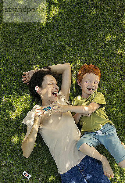 Cheerful mother and son lying on grass and playing with toys in garden