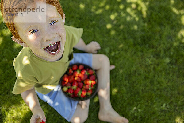 Cheerful boy with mouth stained with strawberries sitting in garden