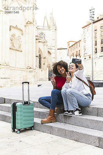 Happy friends making peace sign and taking selfie through smart phone sitting on steps at street