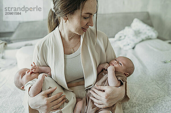 Smiling mother carrying newborn twins and sitting on bed at home