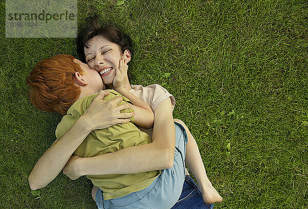 Redhead boy kissing mother lying on grass in garden