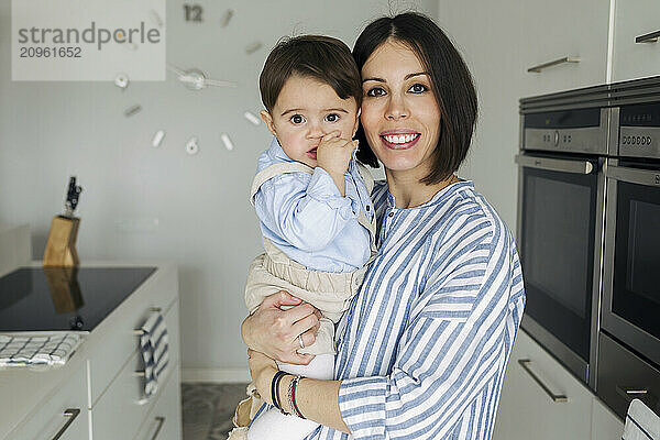 Beautiful woman carrying baby boy and standing in kitchen at home