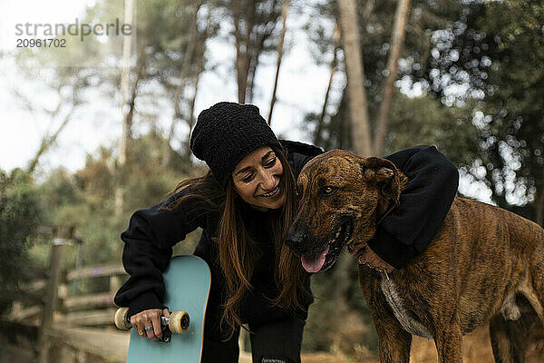 Happy woman holding skateboard and petting dog at park