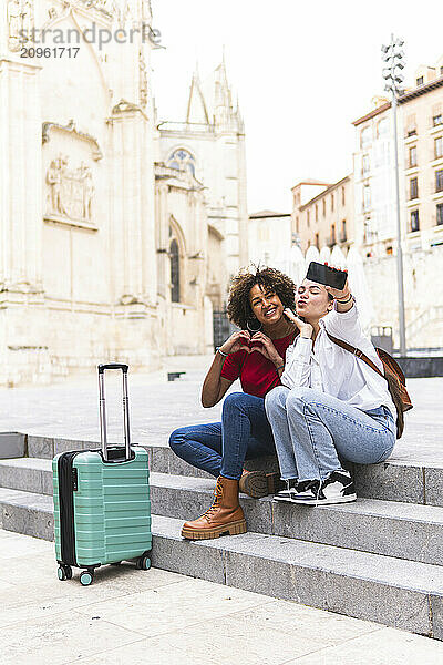 Happy tourist friends gesturing and taking selfie through smart phone sitting on steps at street