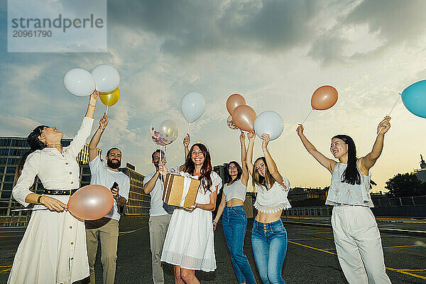 Cheerful friends celebrating woman's birthday with balloons and gift under cloudy sky at sunset