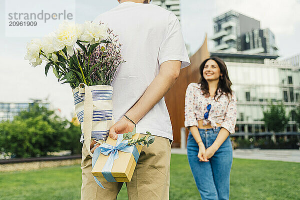 Man hiding gift box and bouquet of flowers in front of woman