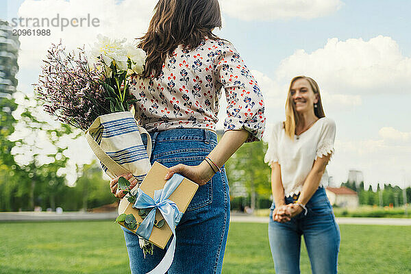 Woman hiding gift box and bouquet of flowers in front of friend