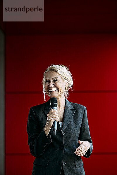 Happy businesswoman holding microphone standing near red wall