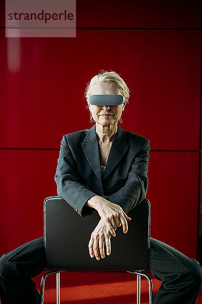 Businesswoman wearing virtual reality headset sitting on chair near red wall