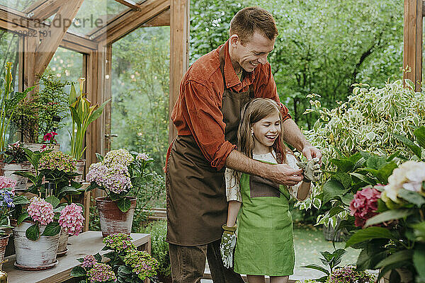 Cheerful girl wearing gardening gloves with help of father at plant nursery