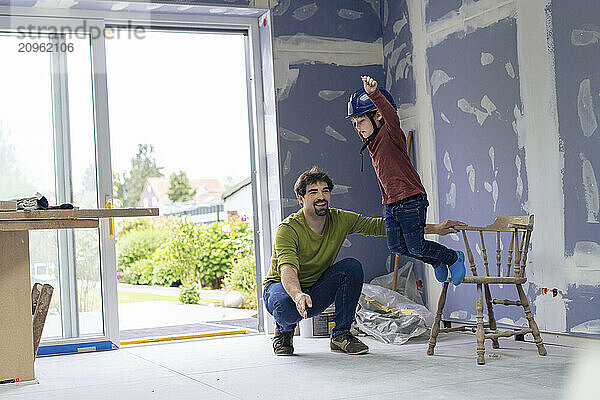 Man crouching near son jumping from chair in new apartment