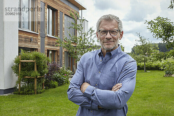 Mature man wearing eyeglasses standing with arms crossed in back yard