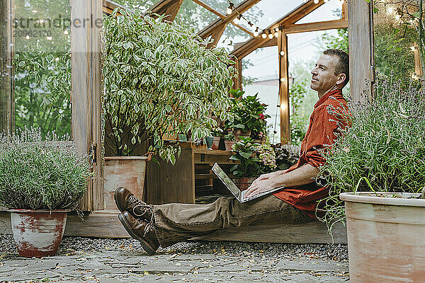 Thoughtful businessman with laptop sitting on doorway of greenhouse