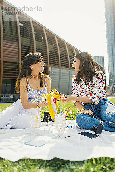 Smiling woman giving gift to friend sitting on picnic blanket