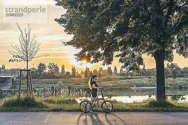 Woman walking with bicycle near tree at sunset