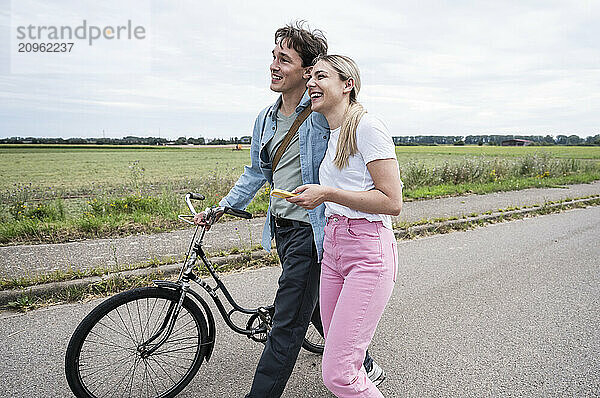 Happy young couple walking with bicycle on road near meadow