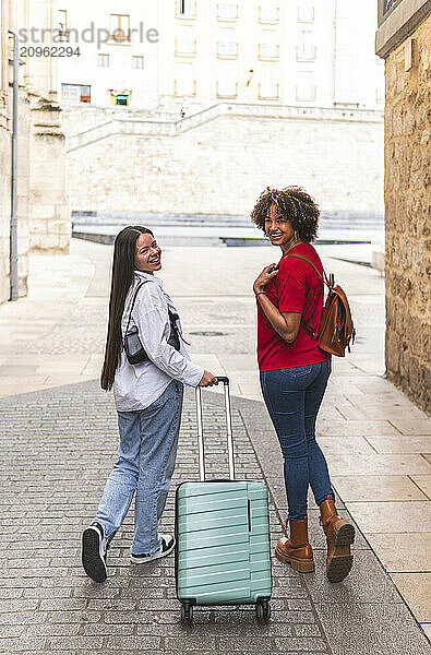 Happy young woman holding suitcase and walking with tourist friend in city