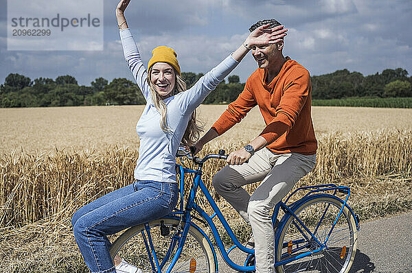 Cheerful woman with arms raised enjoying bicycle ride with man on sunny day