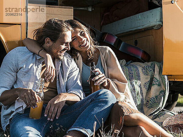 Affectionate young couple with drinks sitting near doorway of van at roadside