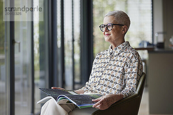 Senior woman sitting in armchair at home with magazine