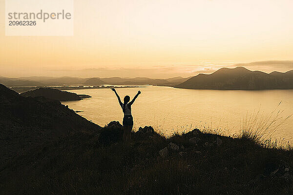 Young woman standing with arms raised at sunrise near river