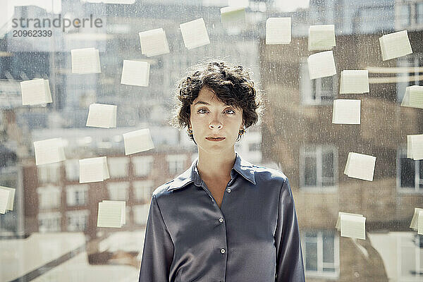 Confident businesswoman standing near glass wall with adhesive notes at office