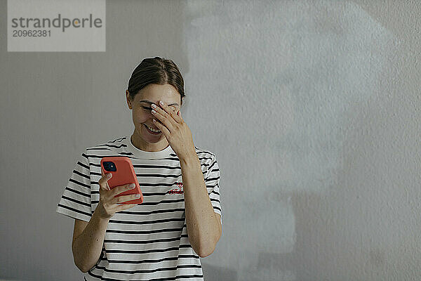 Smiling woman holding smart phone and standing in front of white wall