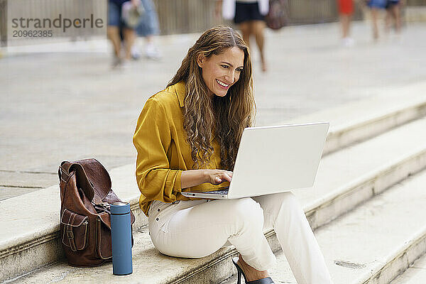 Happy businesswoman using laptop and sitting on steps in city