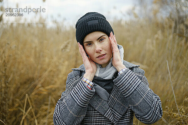 Young woman in warm clothes touching face amidst reeds