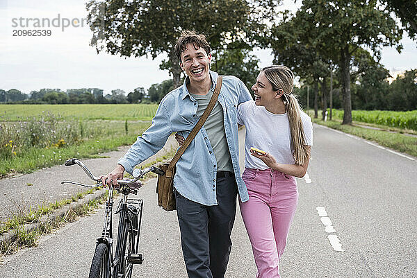 Happy man with arm around girlfriend walking on road amidst meadow