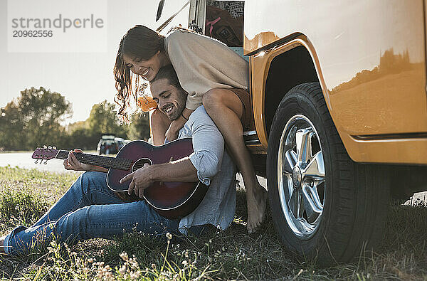 Affectionate couple playing guitar and sitting at doorway of camper van on roadside