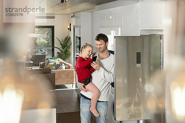 Father and daughter using smart phone standing near refrigerator at home