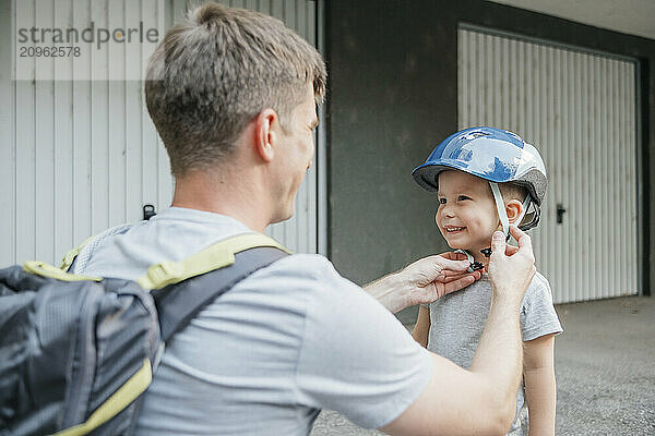 Father fastening safety helmet of son at back yard