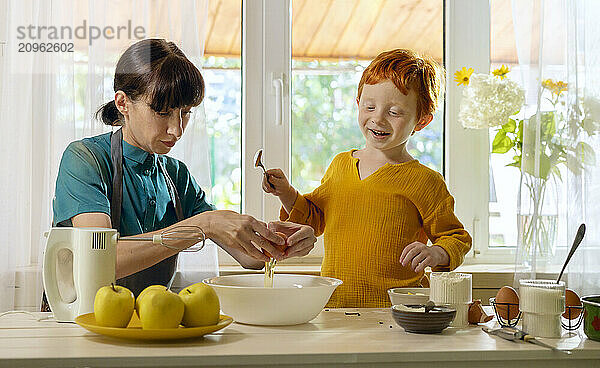 Mother cracking egg into bowl with son standing in kitchen