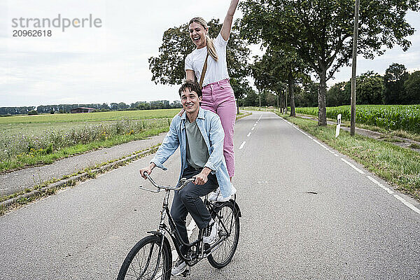 Cheerful woman standing behind boyfriend riding bicycle on road