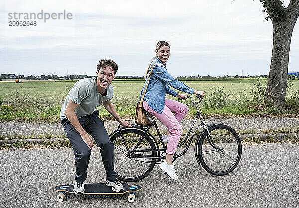 Smiling woman riding bicycle with boyfriend skateboarding on road near meadow