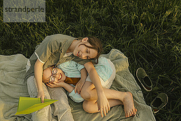 Smiling girl with brother lying on picnic blanket at meadow