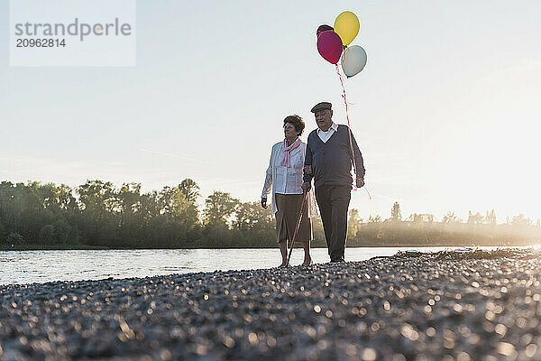 Senior couple walking with balloons near river under sky