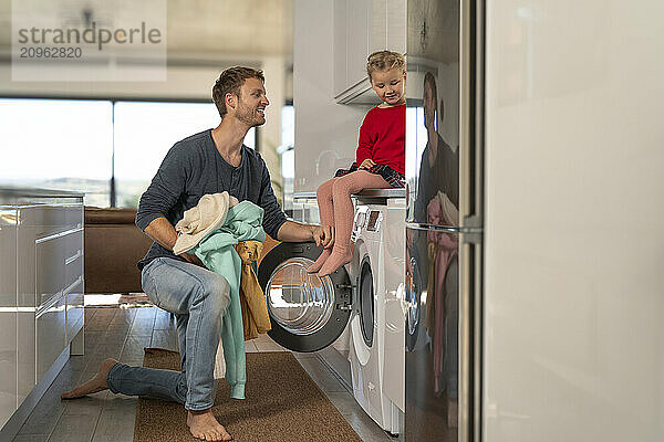 Father loading clothes in washing machine with daughter sitting in kitchen at home