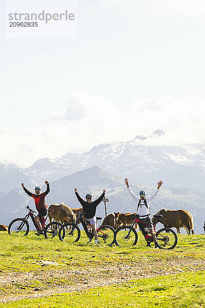 Friends with arms raised standing near bicycle amidst horses on mountain