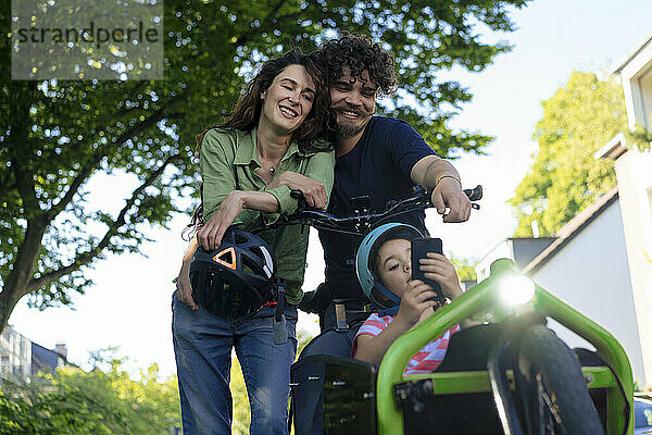 Happy mother and father standing with daughter using smart phone sitting in cargo bike