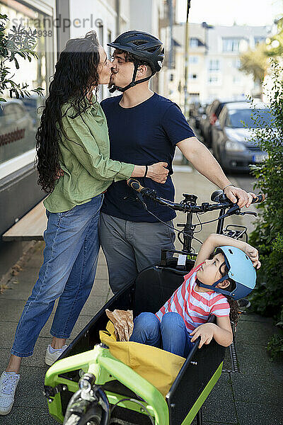 Romantic couple kissing and standing with daughter sitting in cargo bike at street