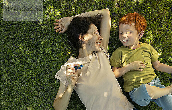 Smiling mother and son lying on grass and playing with toys in garden